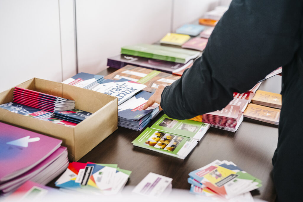 Auf einem Tisch sind unterschiedliche bunte Publikationen zu sehen, an der rechten Bildseite ist der Oberkörper einer Person angeschnitten, deren Hand nach einer der Publikationen greift.