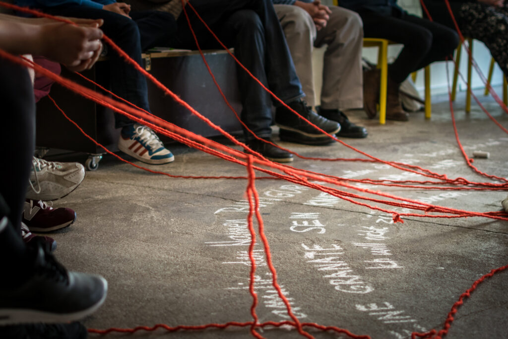 von der unteren linken Ecke des Bildes bis zur oberen rechten Ecke sind die Beine von Menschen, die in einem Kreis auf unterschiedlichen Sitzgelegenheiten sitzen, zu sehen. Am linken Seitenrand des Bildes sieht man Hände, die rote Wollfäden in der Hand halten, auch die anderen Menschen im Kreis halten diese in der Hand, sodass sich zum rechten Seitenrand des Bildes ein Netz aus Fäden entspinnt. Der Boden ist aus grauem Beton, innerhalb des Kreises stehen in weißen Druckbuchstaben Wörter, die nicht klar zu lesen sind.